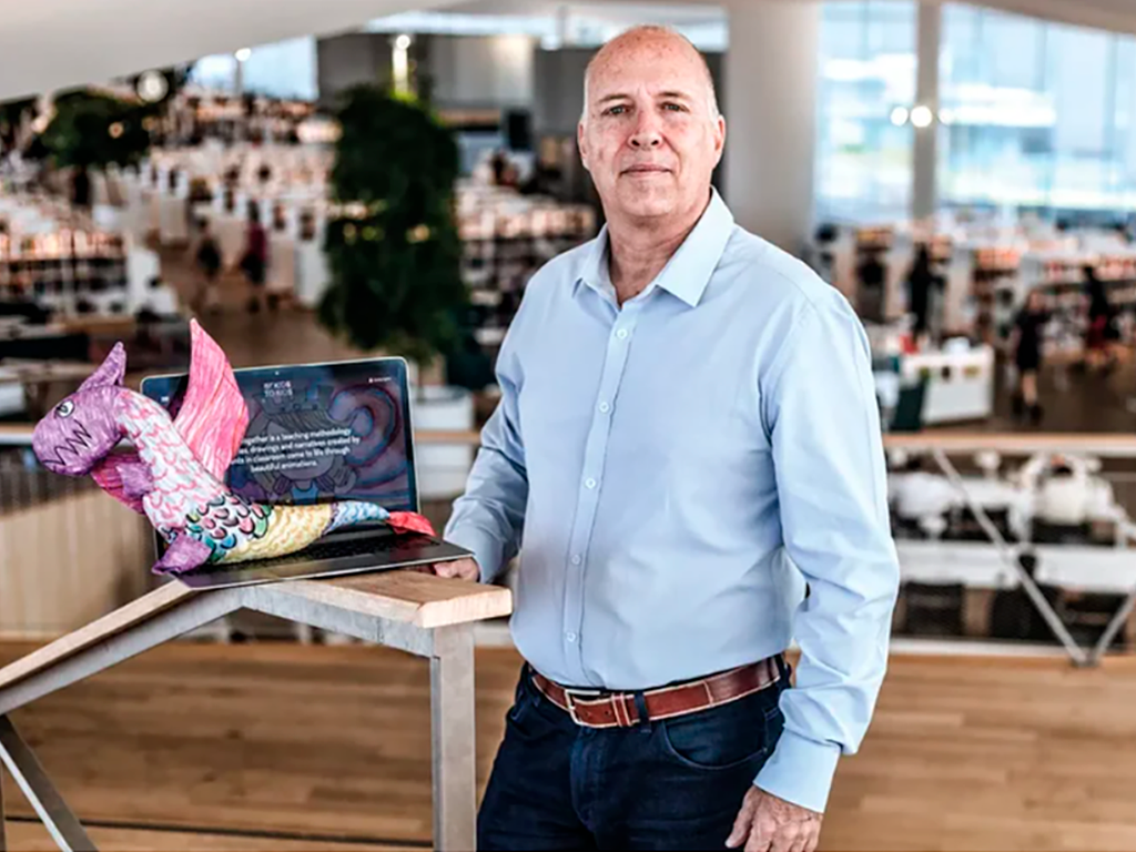 Gilberto Barroso, CEO da DCPC, aparece em primeiro plano. À sua direita, um notebook aberto repousa sobre um corrimão, sugerindo que ele esteja no nível de um mezanino. Ao fundo, observa-se um espaço amplo e iluminado, correspondente ao pavimento inferior. Sobre o notebook, destaca-se um boneco artesanal de tecido em forma de dragão — símbolo das animações 2D desenvolvidas pelo projeto.